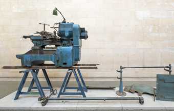 A piece of heavy machinery on a trestle bench on a plinth in a gallery
